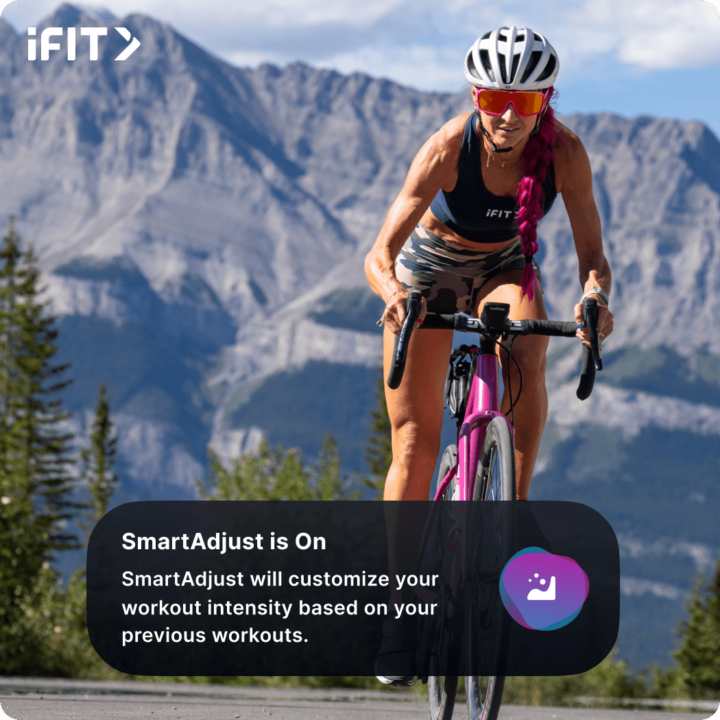 iFIT Trainer Ashley Paulson wearing a white helmet, sunglasses, shorts, and a sports bra riding a pink bike with mountains in the background.