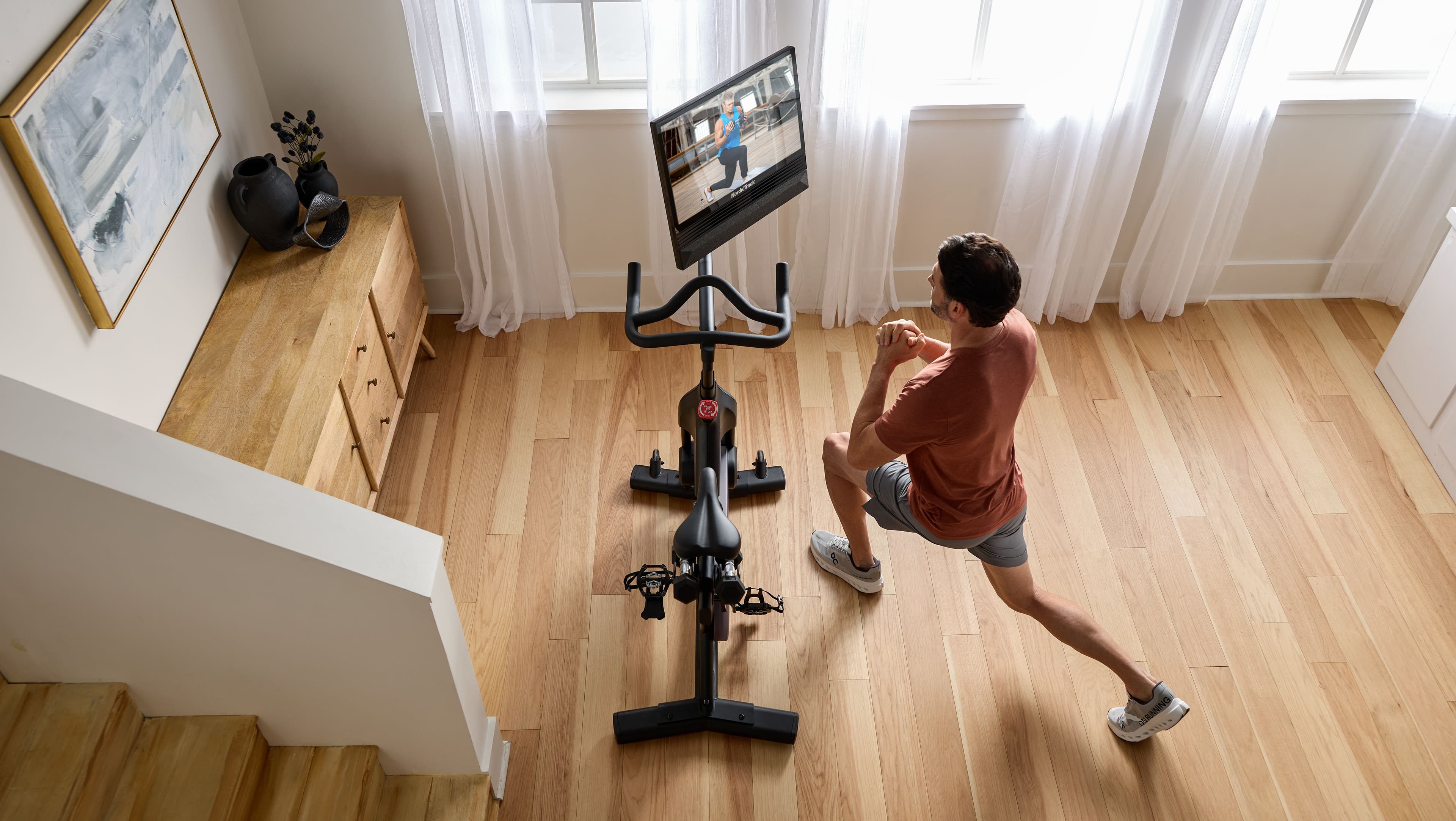 NordicTrack S24 Studio Bike positioned in a compact apartment living room with a man doing a lunge off-bike, following an iFIT workout on the 24-inch touchscreen. Shows real-world apartment footprint and off-bike training capability.
