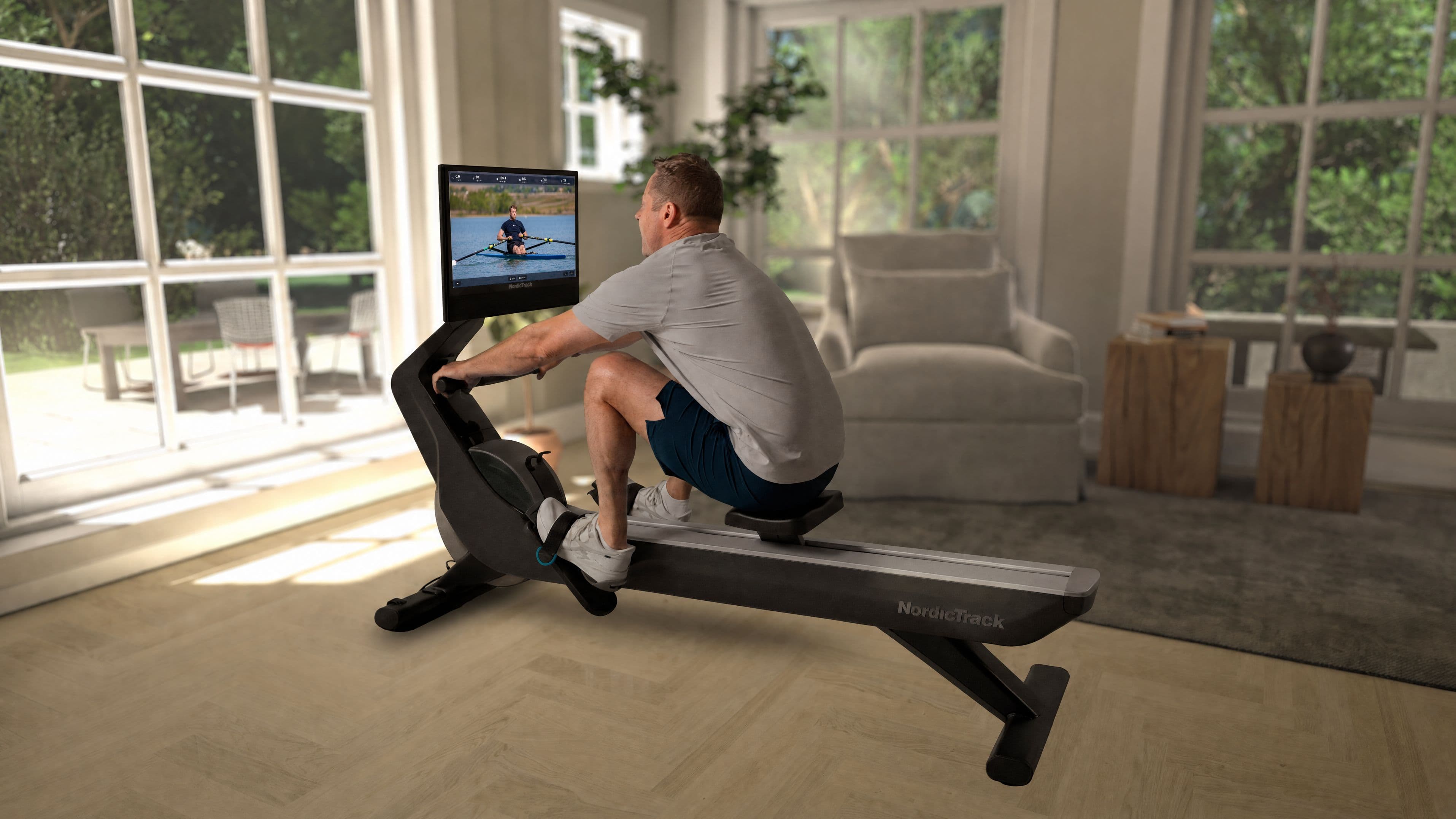 Man using the NordicTrack RW900 rowing machine at home with an iFIT trainer-led workout on the 24-inch touchscreen, in a bright sunroom setting.